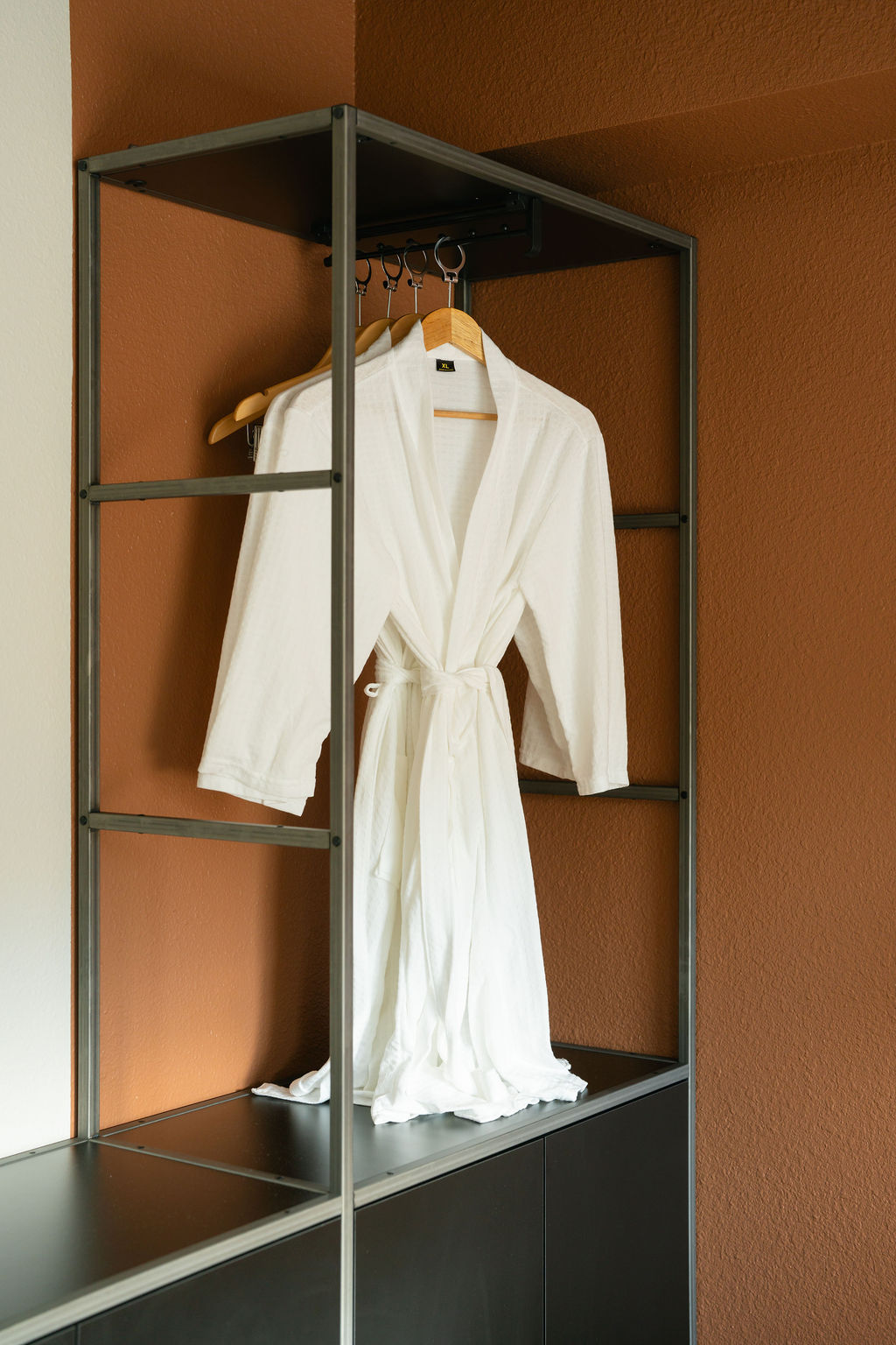 Plush white robe hanging on open wardrobe rack against terracotta accent wall in Outrider Hotel room