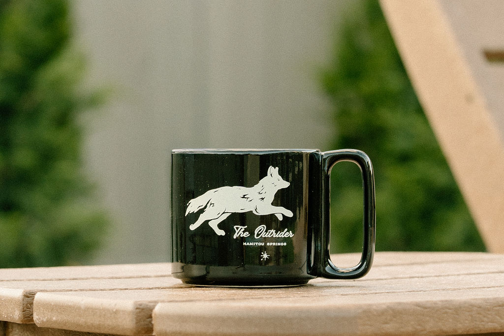 The Outrider Hotel black ceramic mug with wolf logo on outdoor wood table