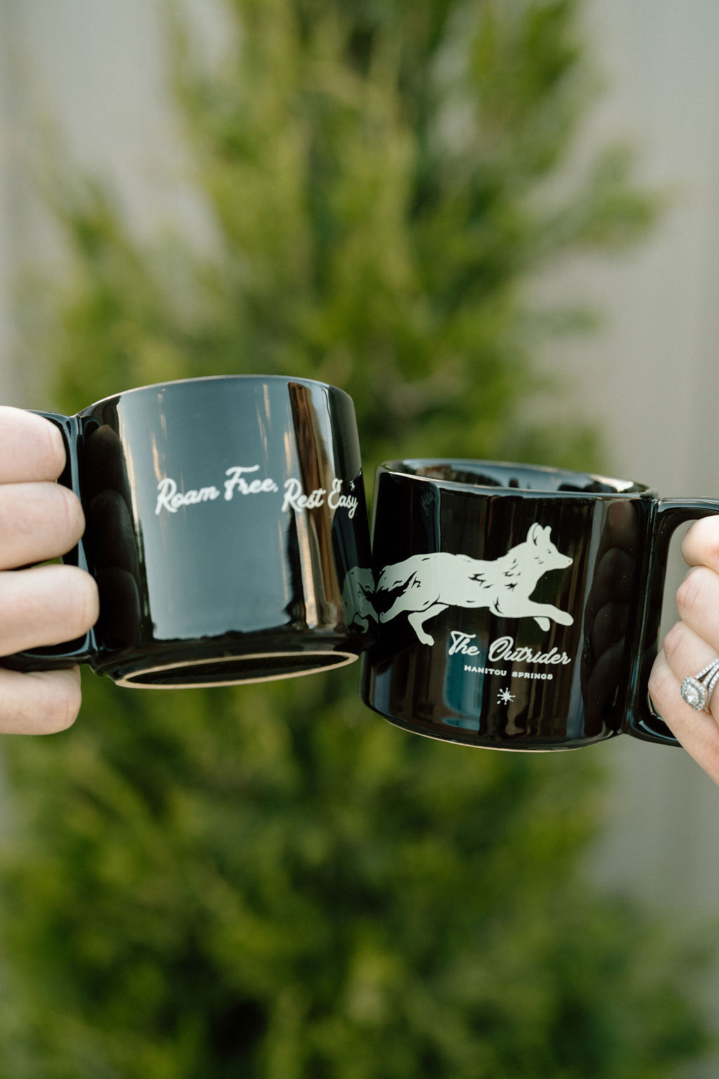 Two Outrider Hotel black mugs reading Roam Free Rest Easy being held up outdoors