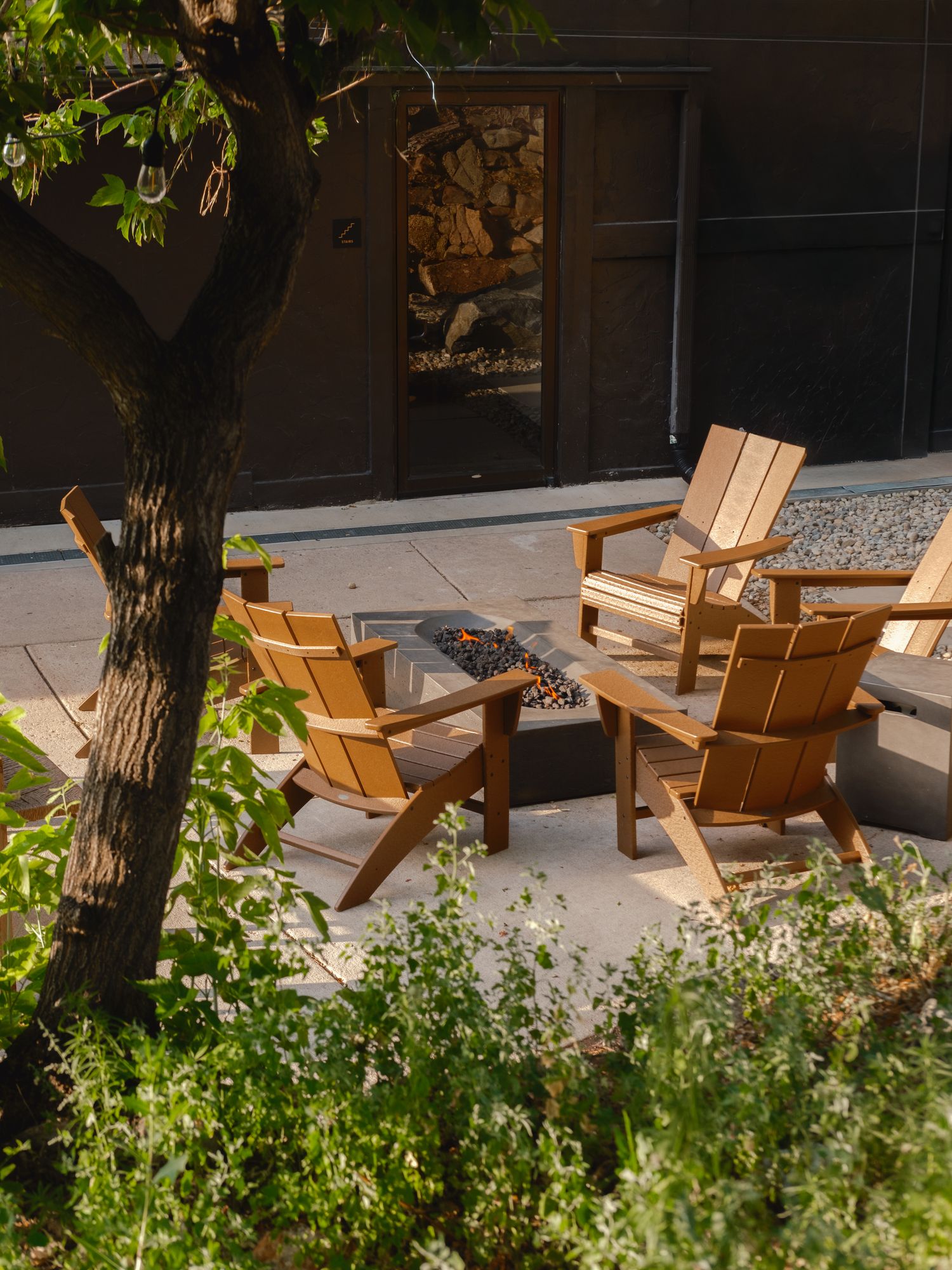 Courtyard fire pit ringed by Adirondack chairs under shade trees at The Outrider Hotel