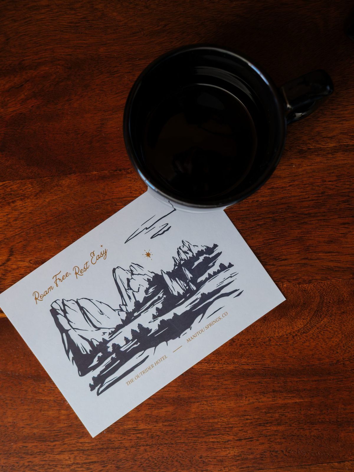 Illustrated Outrider Hotel postcard and black camp mug on a wooden desk